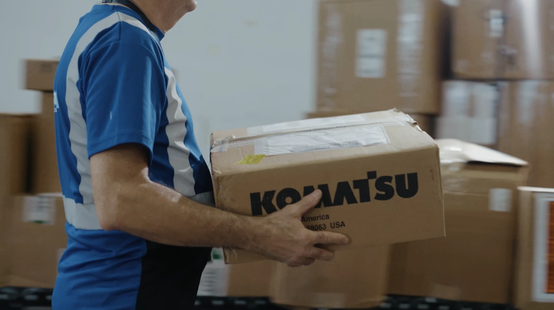 A warehouse worker carrying a Komatsu-labeled box inside a logistics facility, demonstrating careful manual handling and secure cargo processing.